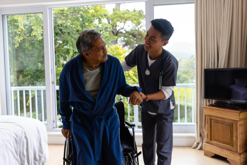 Diverse male doctor helping senior male patient getting up from wheelchair at home. Medical services, check up, home visit, healthcare, wellbeing and senior lifestyle, unaltered.